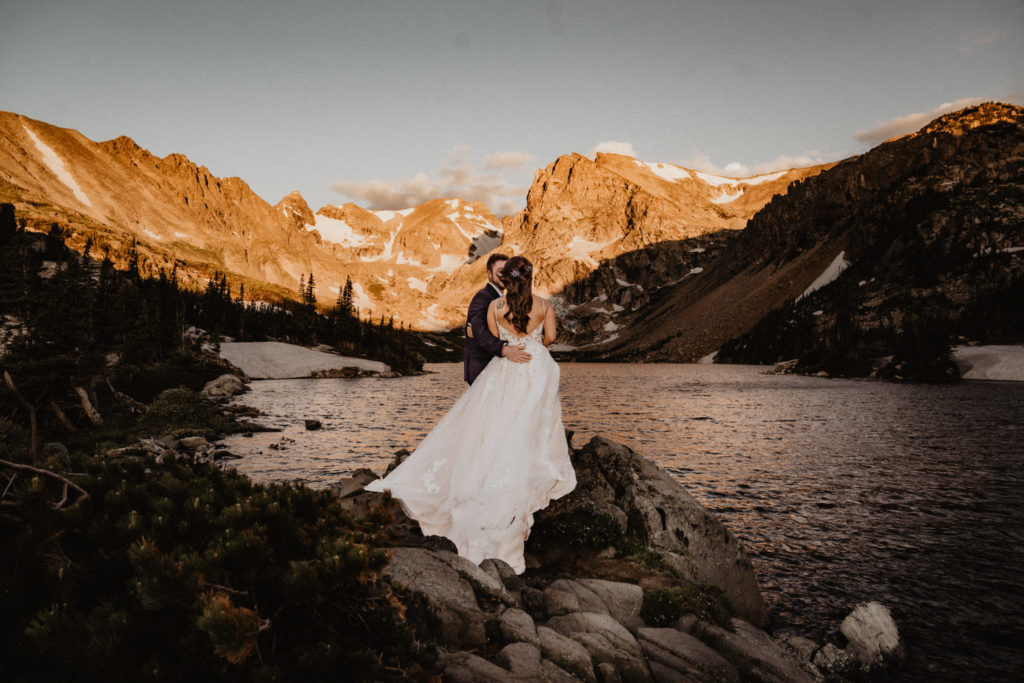 Sunrise Colorado Hiking Elopement where the bride and groom are standing on rocks overlooking a lake surrounded by mountains. Her dress is billowing in the breeze and cascading over the rocks. 