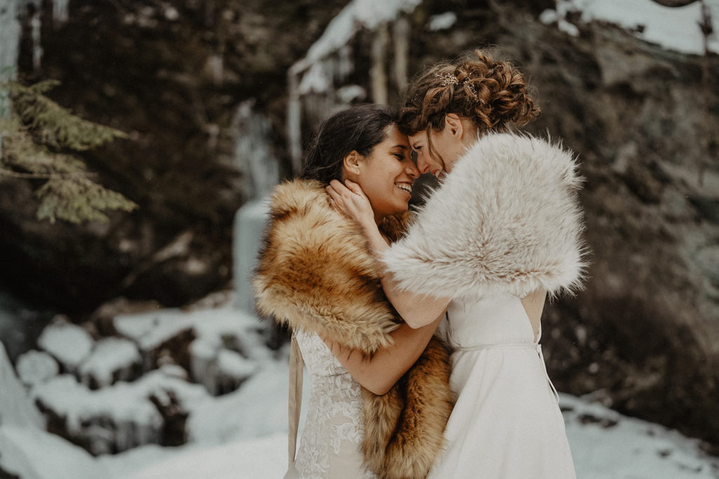 Brides embracing on their elopement day. One wearing a lacy dress with a brown faux fur shawl, the other in a silky open back dress and a white faux fur shawl in a rocky, snowy Stowe VT. 