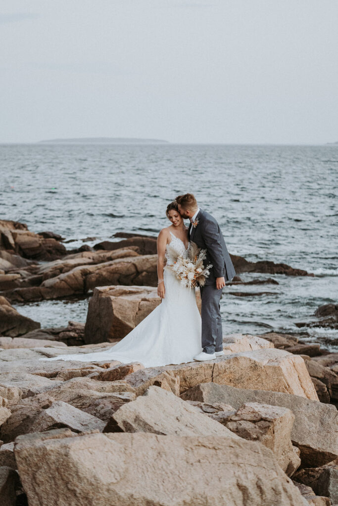 Groom kissing bride on rocky shoreline