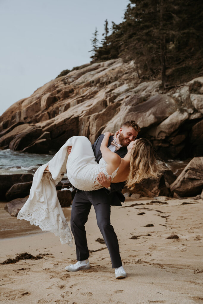 Groom sweeps bride off her feed on the sandy shores surrounded by rocks and water during their elopement. 