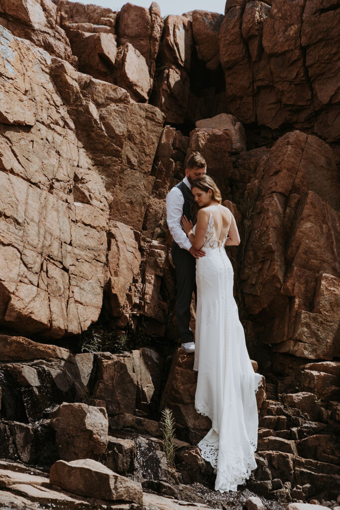 Bride and groom embracing with a rugged rocky background. The lacy train of her open backed elopement dress cascades down the rocks. 