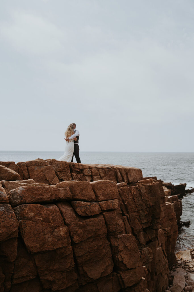 Bride and groom embrace on rocky cliff overlooking the shore during their New England Elopement. 