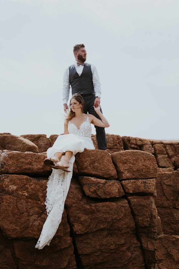 Bride sits on rocky cliff leaning back on the legs of the groom standing behind her during their New England Elopement. 