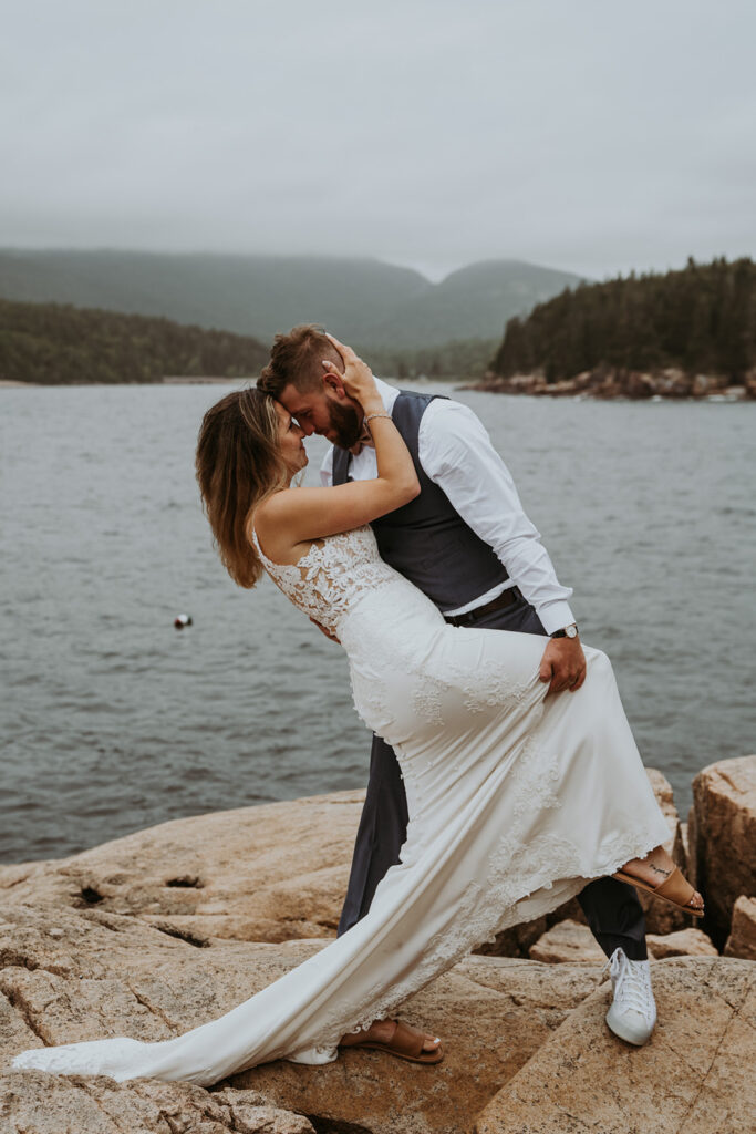 Groom dips bride on rocky shoreline with foggy mountains in the background. Her long lace elopement dress trails over the rocks. 