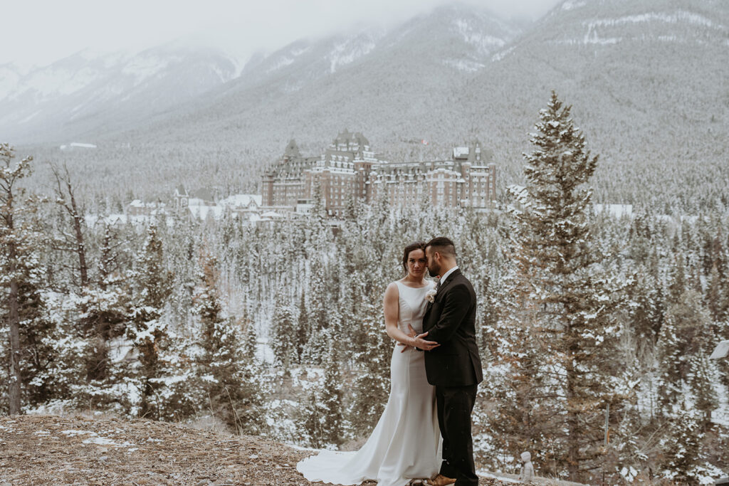 Couple embraces in a mountain top clearing during their Banff National Park Elopement with snow covered trees, peaks and hotel in the background