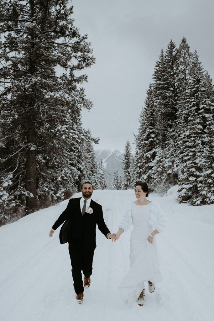 Bride and groom run hand in hand toward camera, with towering snow covered trees in the background celebrating their Banff National Park Elopement. 