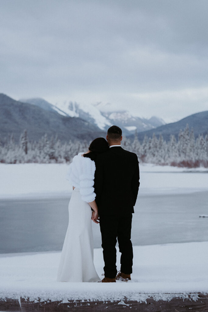 Bride rests her head on grooms shoulder as they look out at one of the icy lakes in Banff National Park during their adventure elopement. 