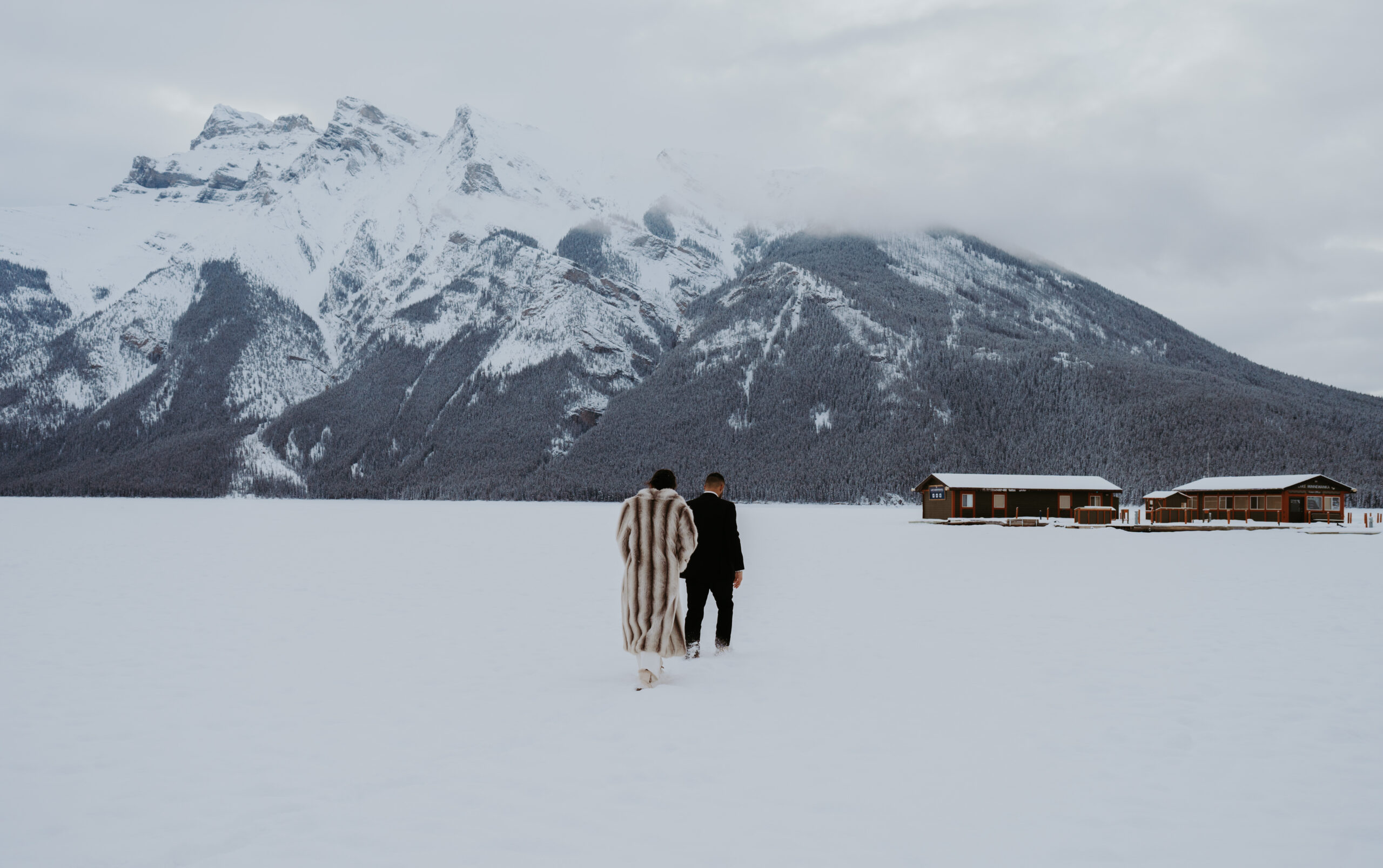 Bride and groom walking on the ice of lake minnewanka towards the boat house with the rocky mountains of Banff National Park in the background