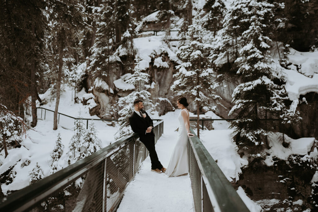 Bride and groom stand opposite each other on a snow covered bridge in the rocky forests of Banff National Park during their adventure elopement. 