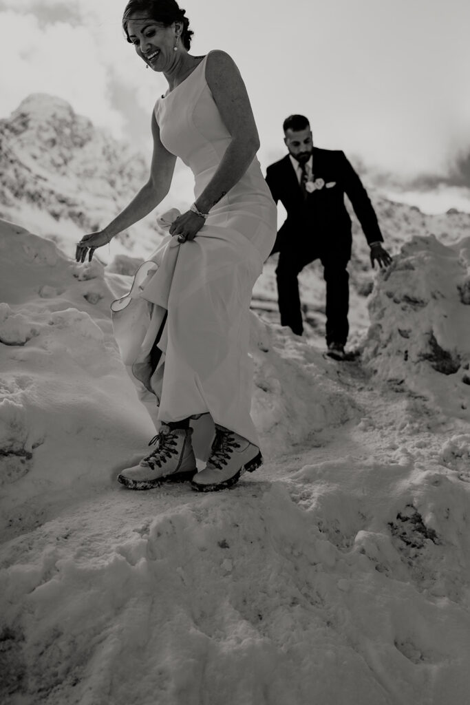 Bride in wedding dress and hiking boots, followed by the groom, hikes down a rocky section in Banff National Park during their adventure elopement. 