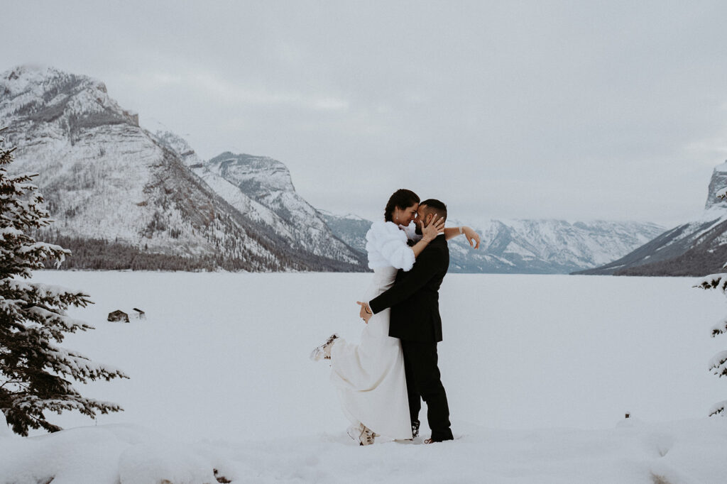 Groom holds bride in front of one of the ice covered lakes of Banff National Park at their adventure elopement. 