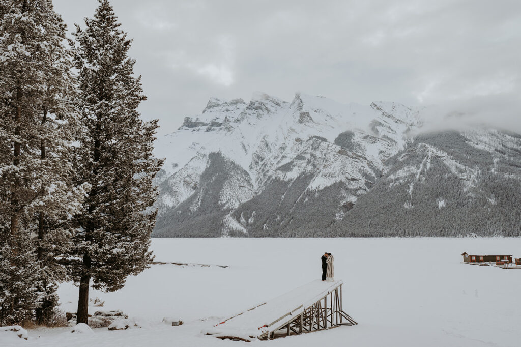 Bride and groom stand together in distance on a ramp overlooking a snow covered field. Tall, snowy trees and mountains tower around them in Banff National Park. 