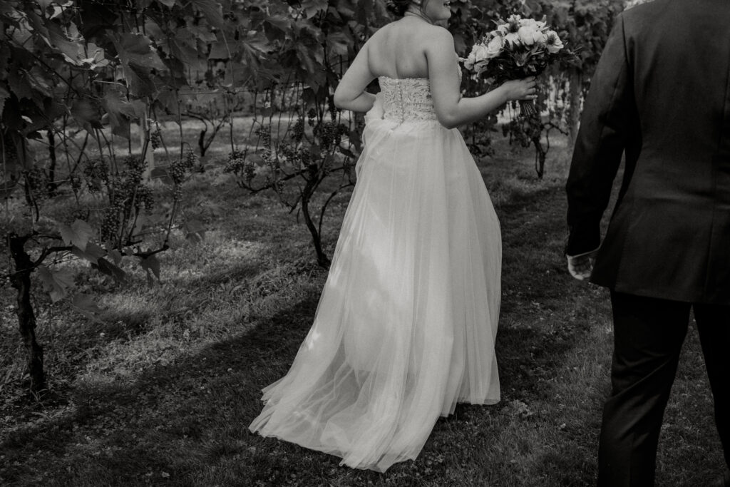 Bride holds her wedding dress as she walks away with her groom by her side through a vineyard at Zorvino Vineyards.