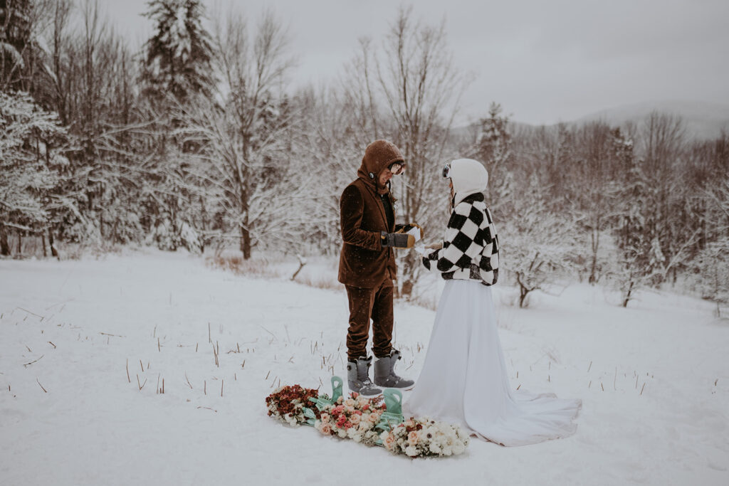 Couple elopes slope-side with floral snowboard in foreground, white dress billowing in the snow. 