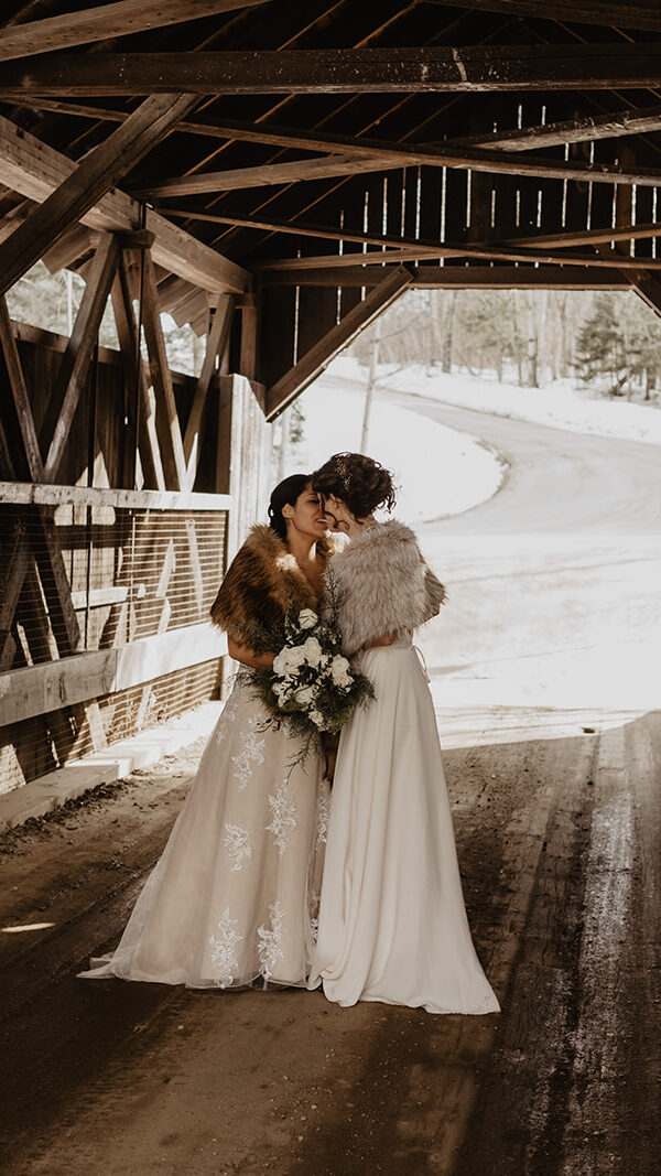 Two brides wearing gowns, and faux fur shawls, kiss under the cover of a wooden bridge during their New England Elopement. 