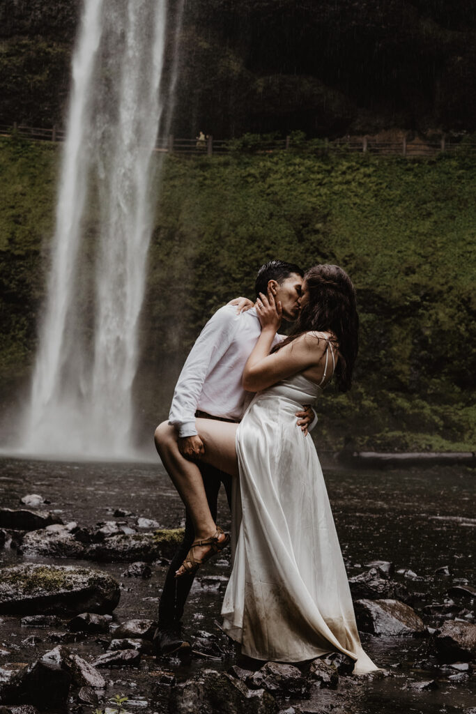 Bride and groom kiss passionately in front of a waterfall and jungle like forrest during their destination elopement.