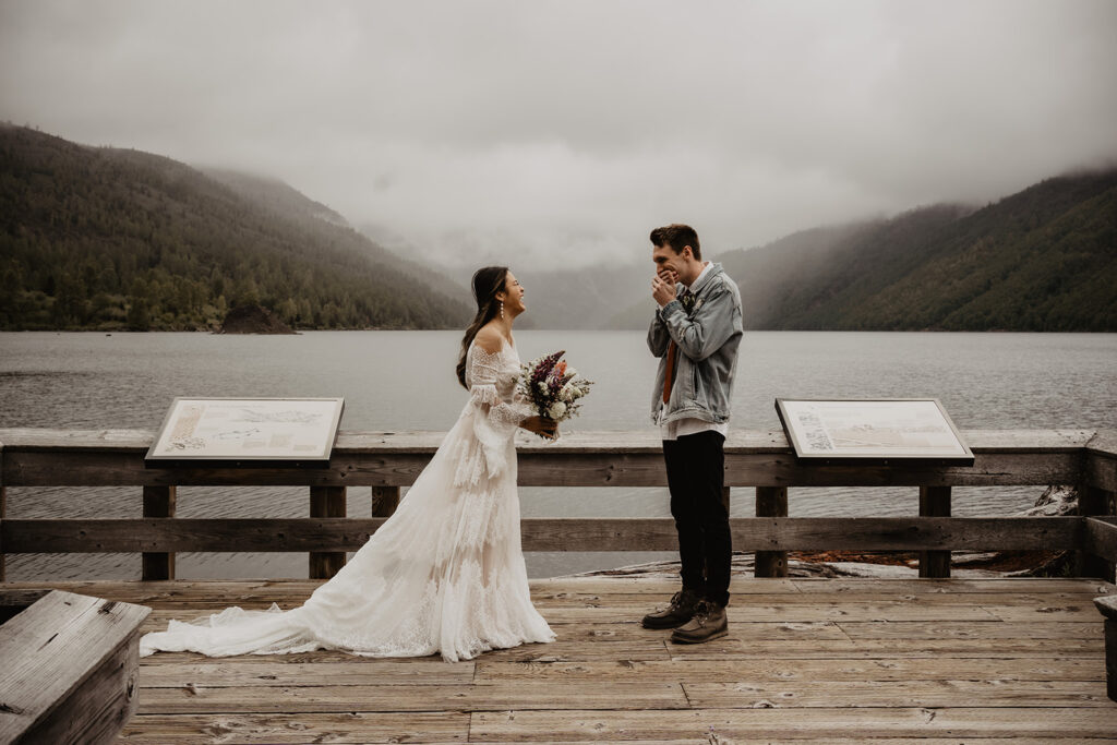 Bride and groom stand facing each other during a first look at their destination elopement with water and fog dipped mountains in the background.