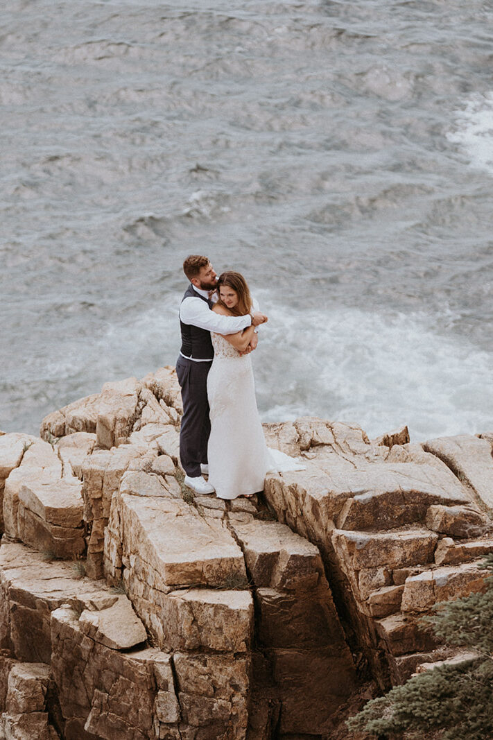 Groom embraces bride on rocky cliff with waves crashing behind them during their New England Elopement. 
