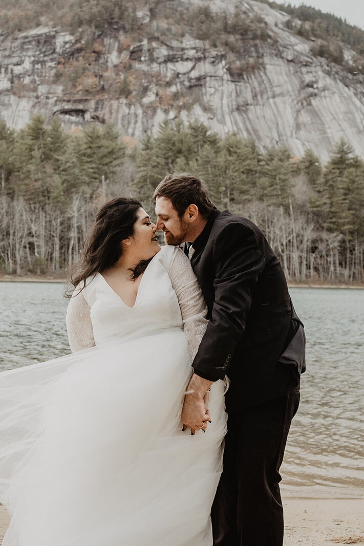 Bride and groom embrace in front of a lake with forrest in the background of their New England Elopement. 