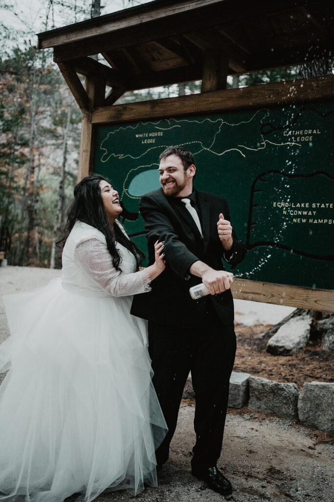 Bride and groom spray champagne in front of state park map during their New England Elopement. 