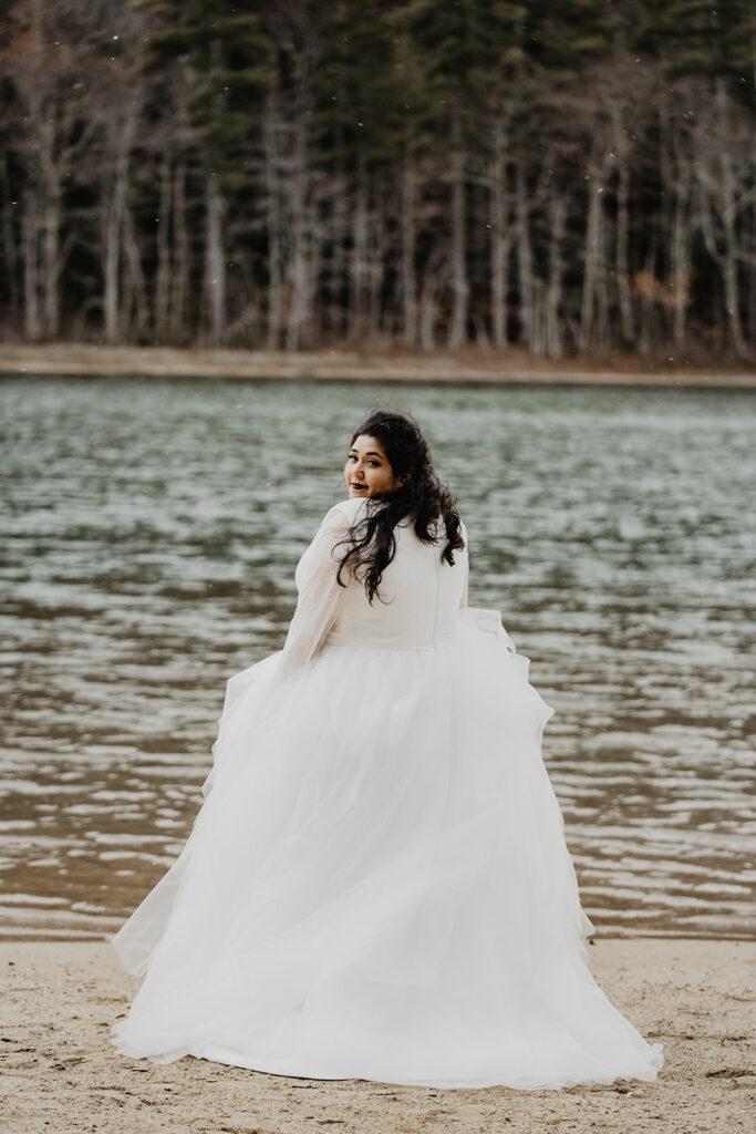 Bride, standing on the waters' edge with a forrest in her background, looks back over her shoulder. 