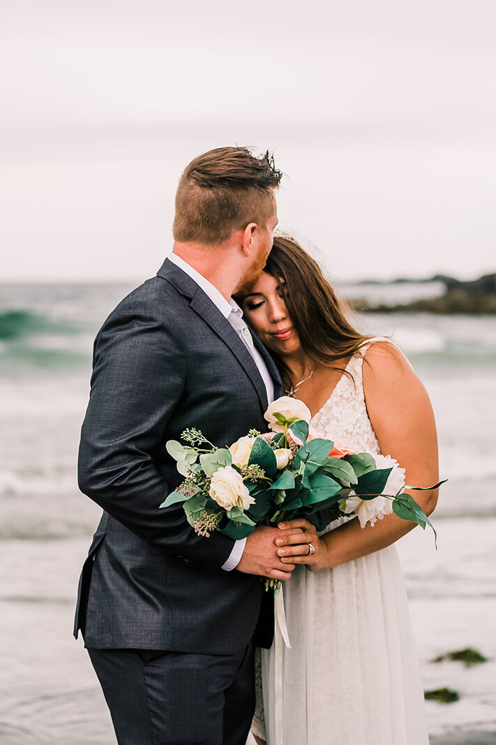 Bride and groom embrace in front of the crashing waves during their New England Elopement. 