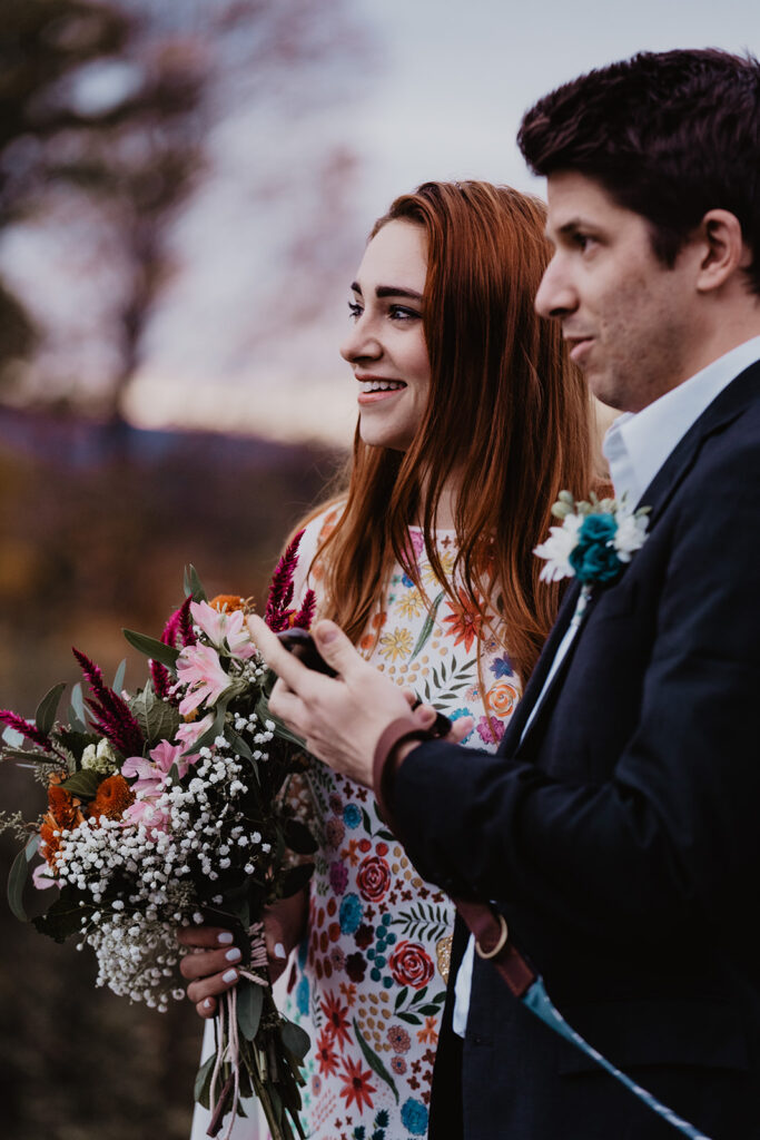 Bride and groom standing side by side. She is holding her colorful bouquet and both are looking away. The sunset tones of purples and red are faded into the background during this New England Elopement. 
