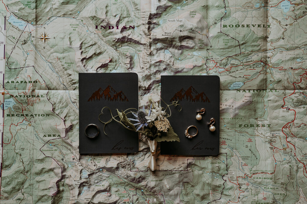 Black vow books, with boutonniere wedding rings and earrings all laid atop a map.  