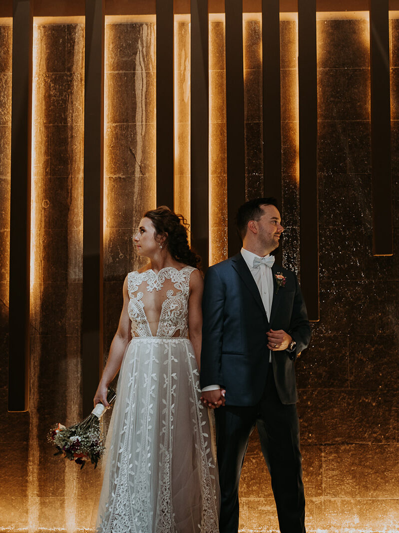 Bride and groom stand next to each other, holding hands and looking in opposite directions against a back lit wall at their venue in Costa Rica. 