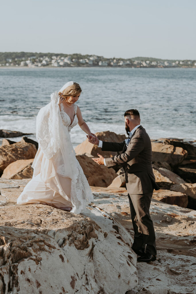 Groom helps bride walk on the rocky shore during their New England Elopement. 