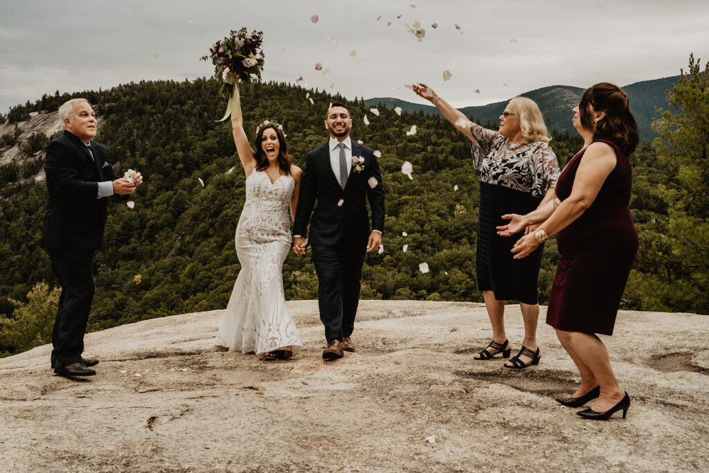 Bride and groom celebrate as guests throw rose petals in the air at their intimate mountain top elopement 