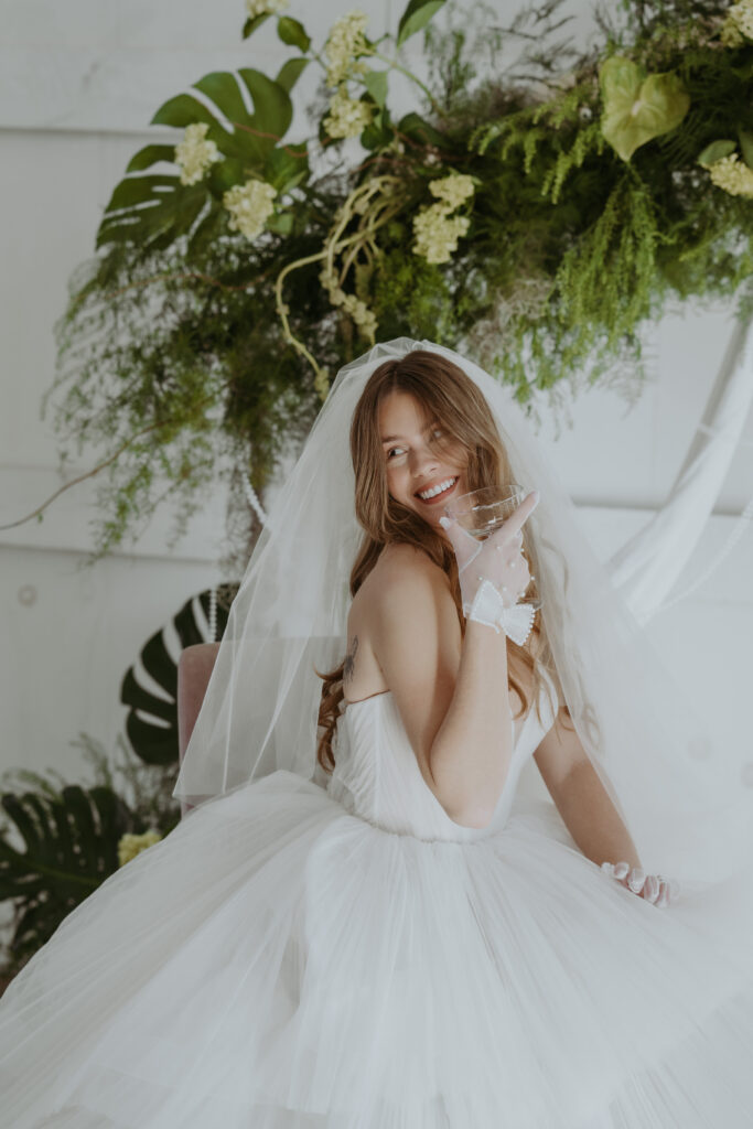 Bride sipping from champagne glass looking behind her in big ballgown at the Greenery wedding venue, shot by Boston Wedding photographer Lexi Foster